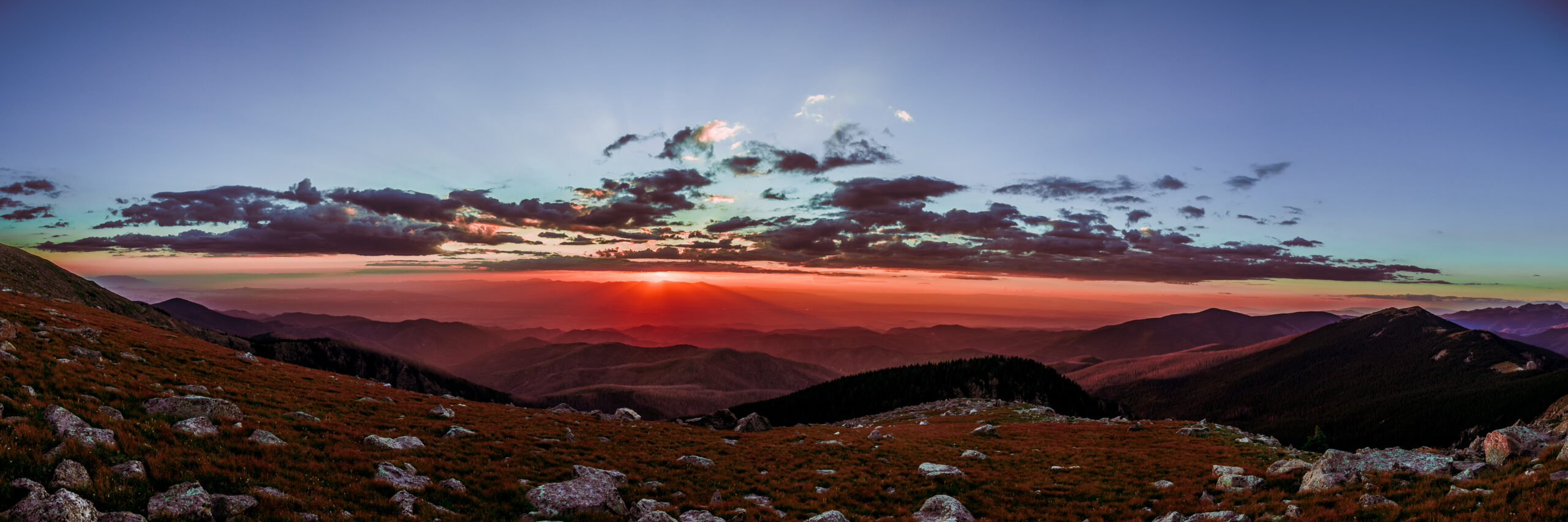 Panoramic sunset view from Santa Fe Baldy showing vibrant red, orange, and purple skies over layered mountain ridges and rocky foreground.